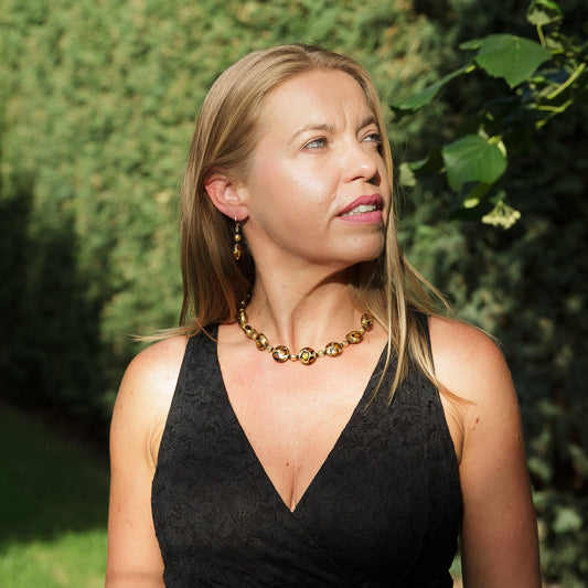 Woman wearing a black dress and gold necklace outdoors with greenery in the background